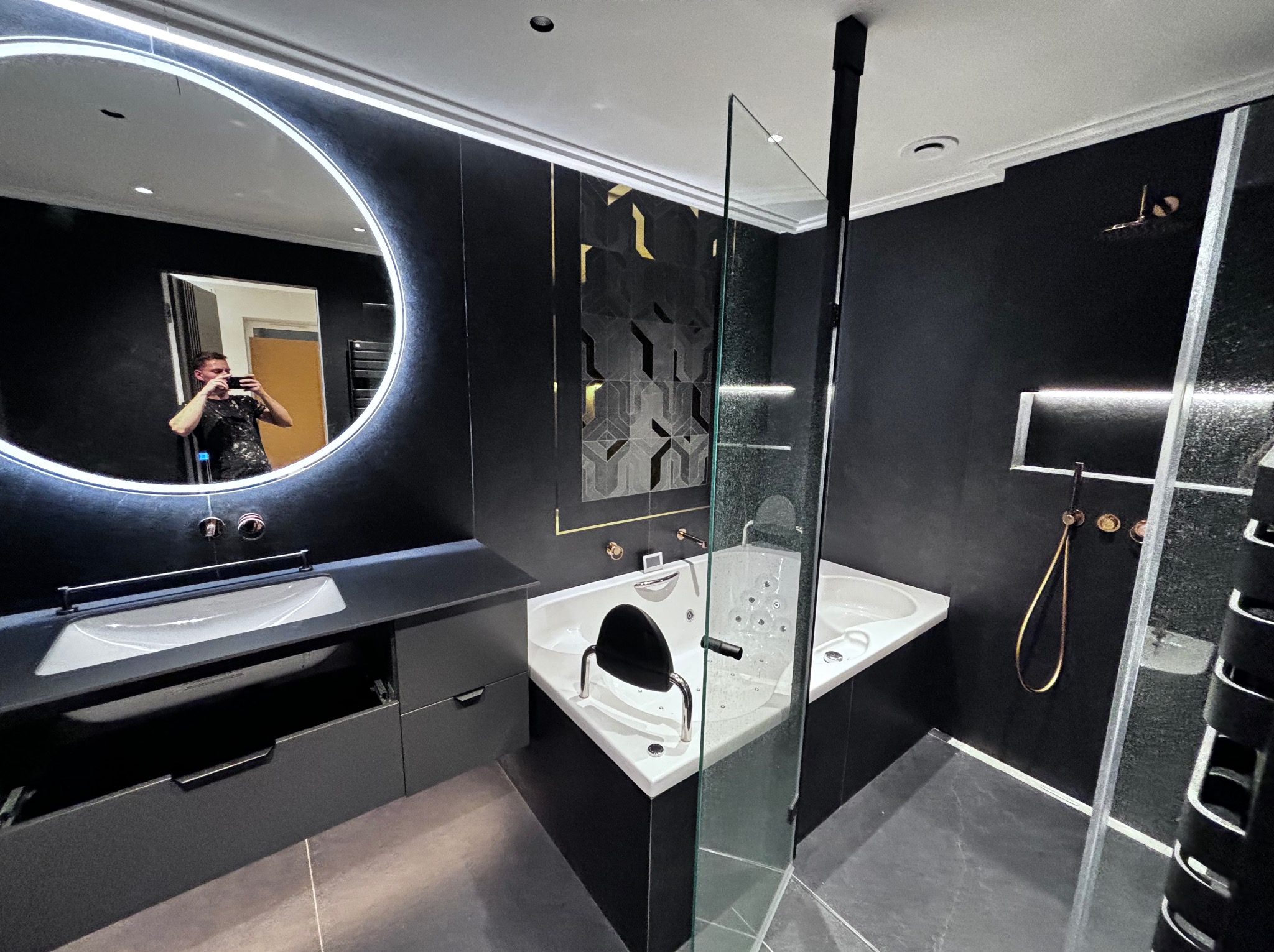 Nowoczesna łazienka z czarnymi ścianami, okrągłym podświetlanym lustrem odbijającym fotografa, białą wanną z hydromasażem i szklaną kabiną prysznicową.