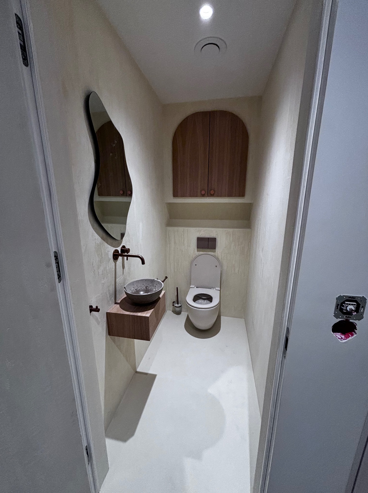 Wąska łazienka z toaletą, umywalką w kształcie misy na drewnianym blacie, asymetrycznym lustrem i szafką nad toaletą, wykończona w jasnych, beżowych odcieniach z akcentami miedzianego koloru.