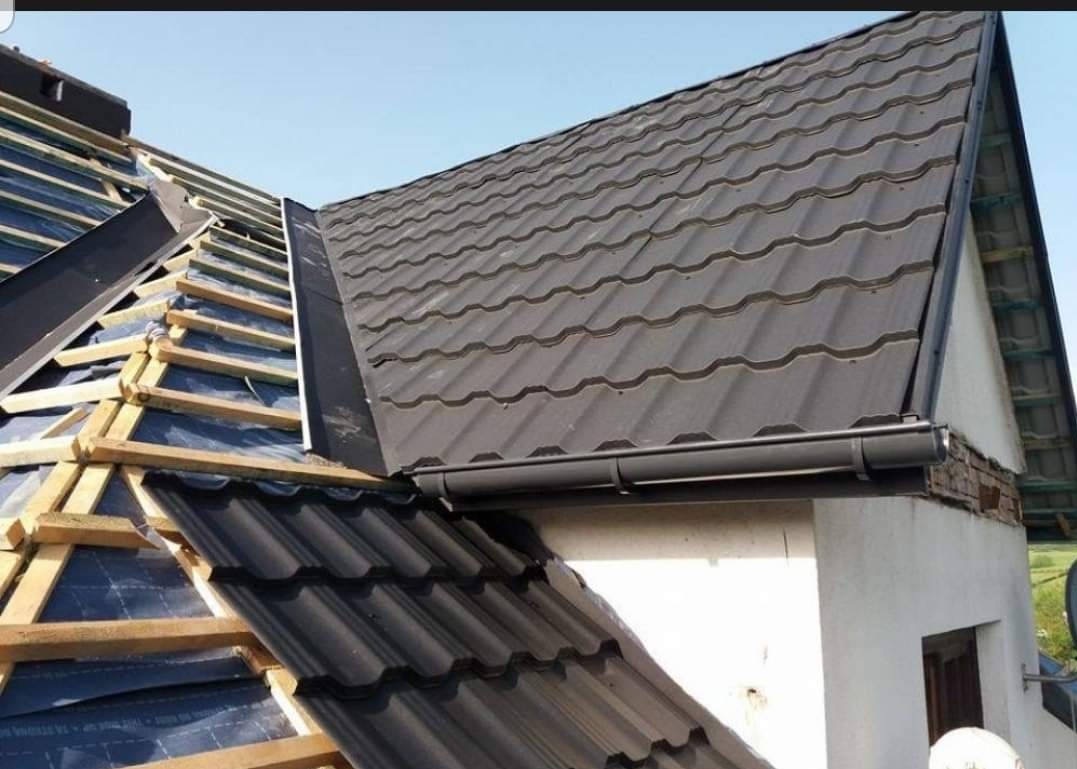 Fragment dachu z częściowo ułożoną, ciemną blachodachówką falistą, widoczne elementy konstrukcji drewnianej i rynna w kolorze grafitowym.