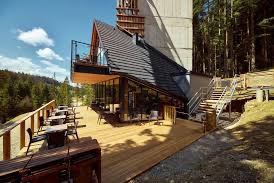 Nowoczesny budynek z drewnianym tarasem i skośnym dachem pokrytym grafitową dachówką, widoczny z perspektywy żaby na tle lasu i błękitnego nieba.