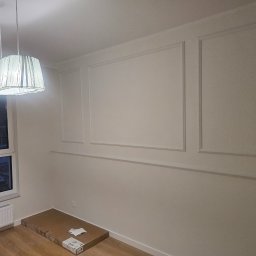 Rafał Erbetowski - Pomalowane na biało wnętrze z dekoracyjnymi listwami ściennymi, widoczny fragment podłogi z panelami, okno z grzejnikiem i lampa wisząca z abażurem, na podłodze karton i ulotki.