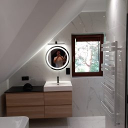 Rafał Erbetowski - Nowoczesna łazienka na poddaszu z marmurowymi płytkami, okrągłym lustrem z podświetleniem LED, drewnianą szafką pod umywalką i grzejnikiem drabinkowym; odbicie fotografa w lustrze.