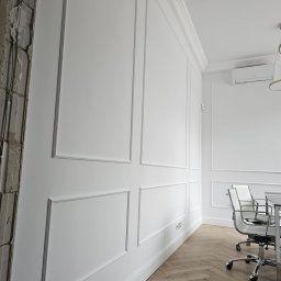 Rafał Erbetowski - Biała ściana z dekoracyjnymi panelami i listwami przypodłogowymi, fragment surowego muru z lewej strony, parkiet w jodełkę, biurowe krzesła w tle.