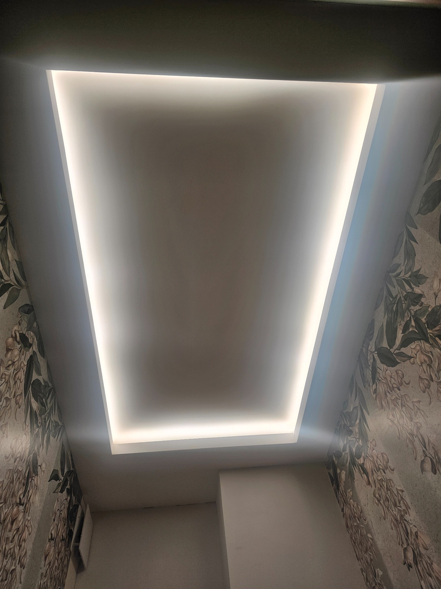 Podwieszany sufit z oświetleniem LED w kształcie prostokąta, otoczony ścianami z tapetą w roślinny wzór.