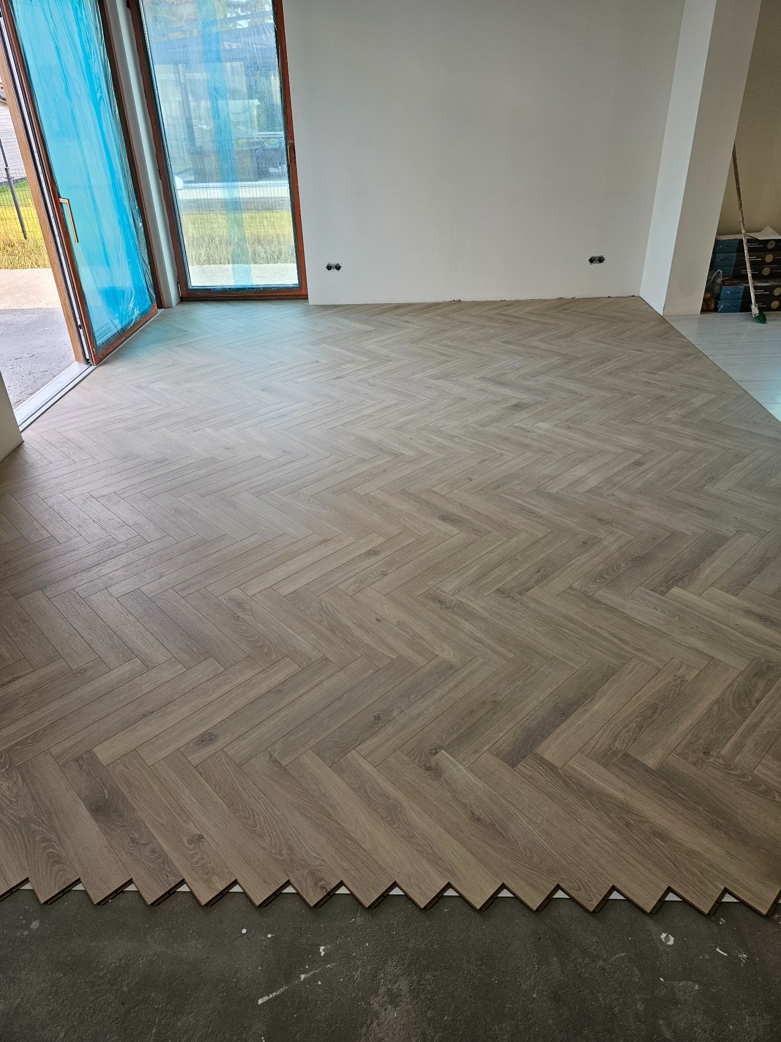 Układanie parkietu w jodełkę w nowym mieszkaniu, widoczne niedokończone krawędzie przy ścianie i osłonięte folią okno balkonowe.