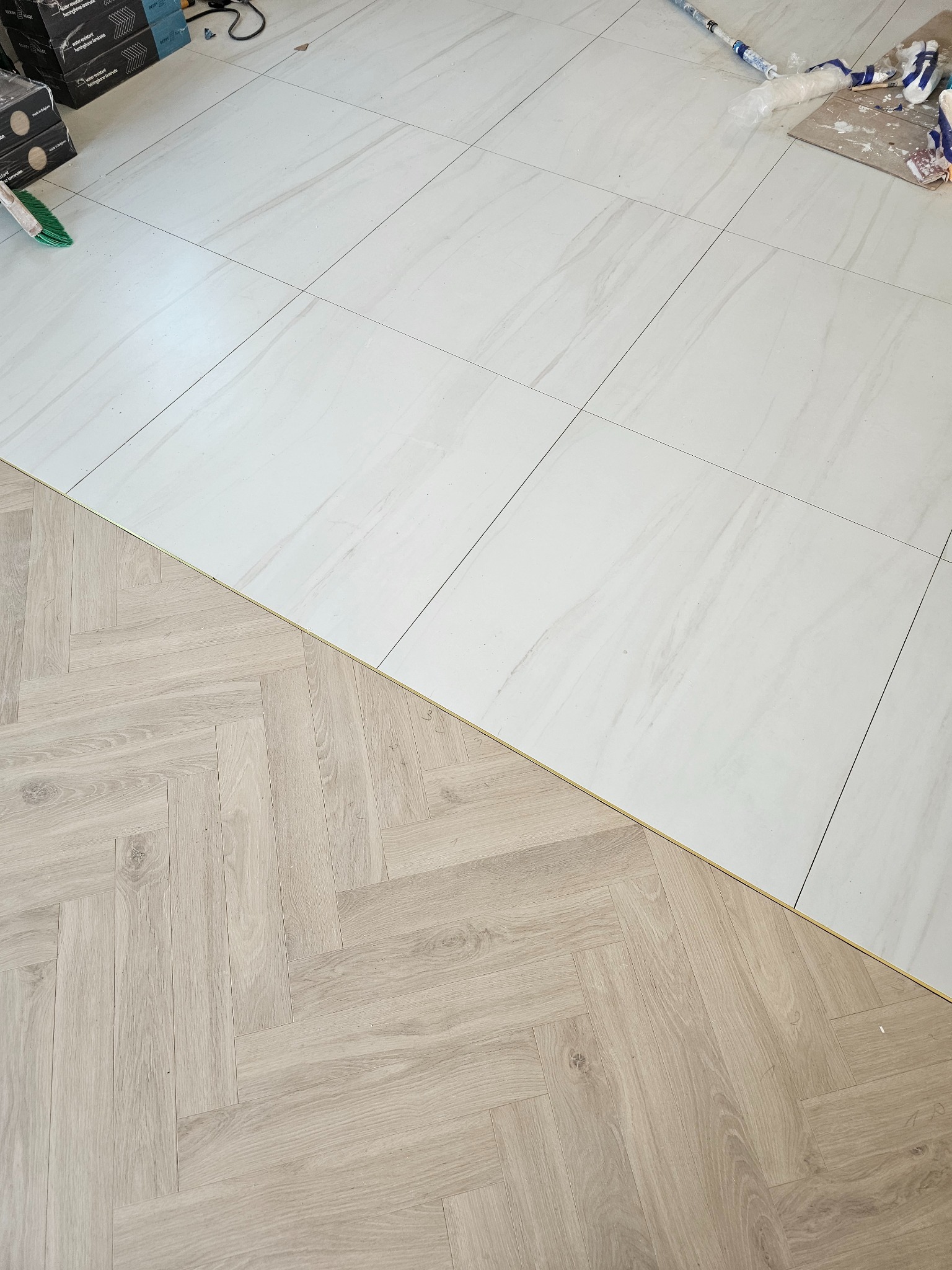 Połączenie jasnych, marmurkowych płytek podłogowych i paneli ułożonych w jodełkę, widoczne narzędzia i materiały budowlane w tle.