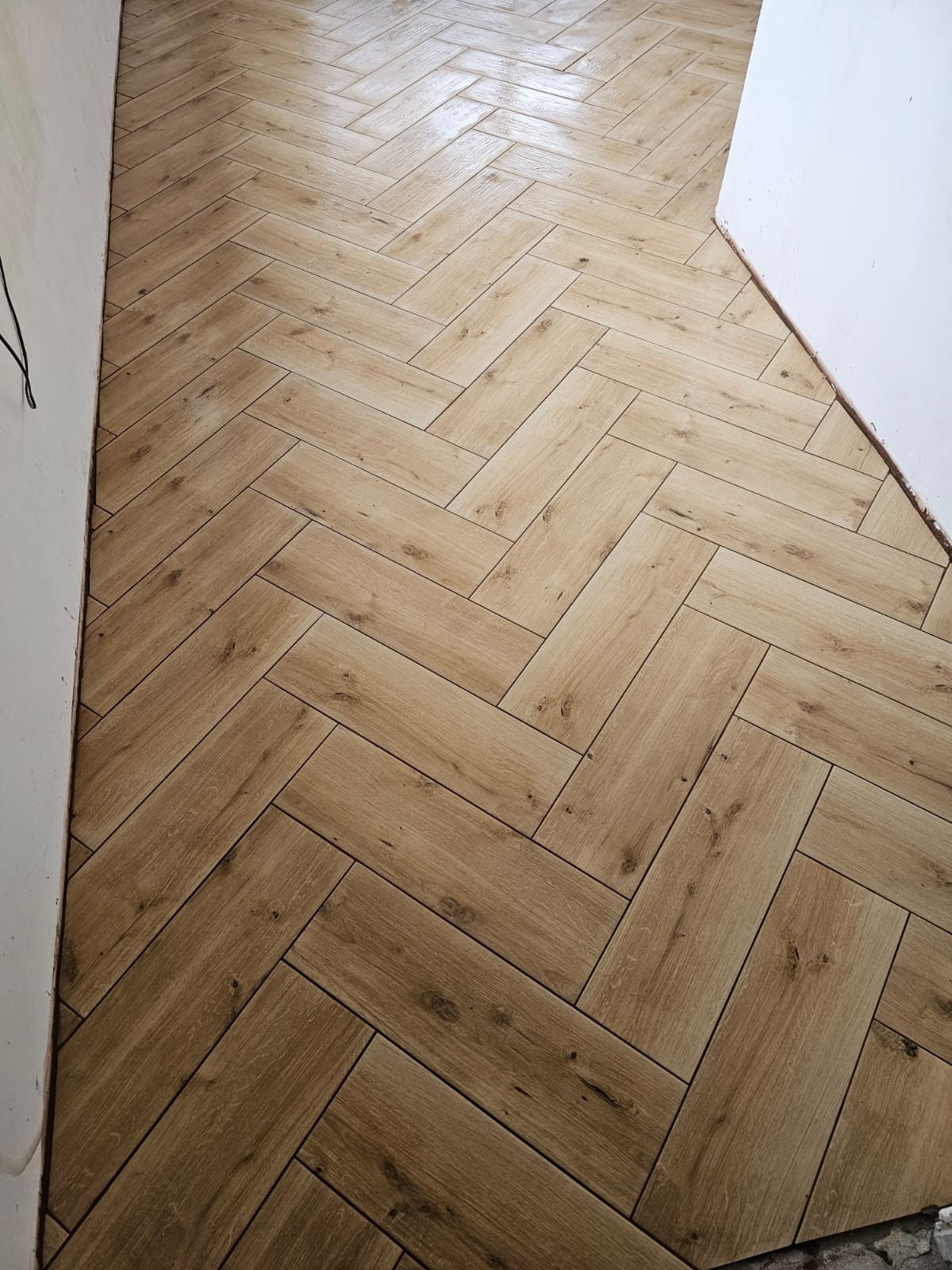 Ułożona pod kątem jodełki, błyszcząca podłoga z płytek imitujących drewno, widok z góry na fragment pomieszczenia z białymi ścianami.