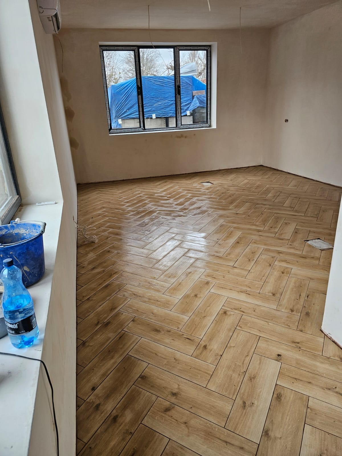 Ułożona, błyszcząca podłoga z płytek imitujących drewno w jodełkę w pokoju w trakcie remontu; widoczne niedokończone ściany, okno z widokiem na budynek przykryty niebieską plandeką, wiadro...