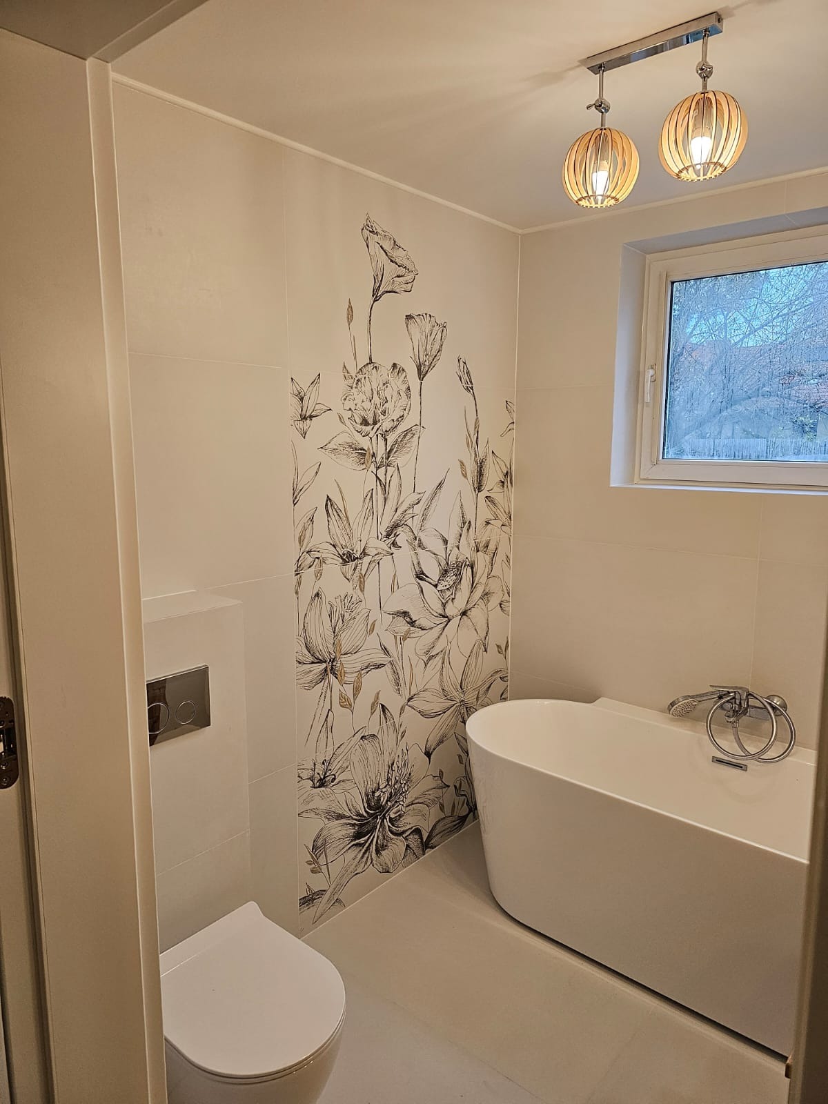 Nowoczesna łazienka z wolnostojącą wanną, toaletą podwieszaną i ścianą wyłożoną płytkami z motywem kwiatowym w odcieniach szarości i beżu, oświetlona drewnianymi lampami wiszącymi.