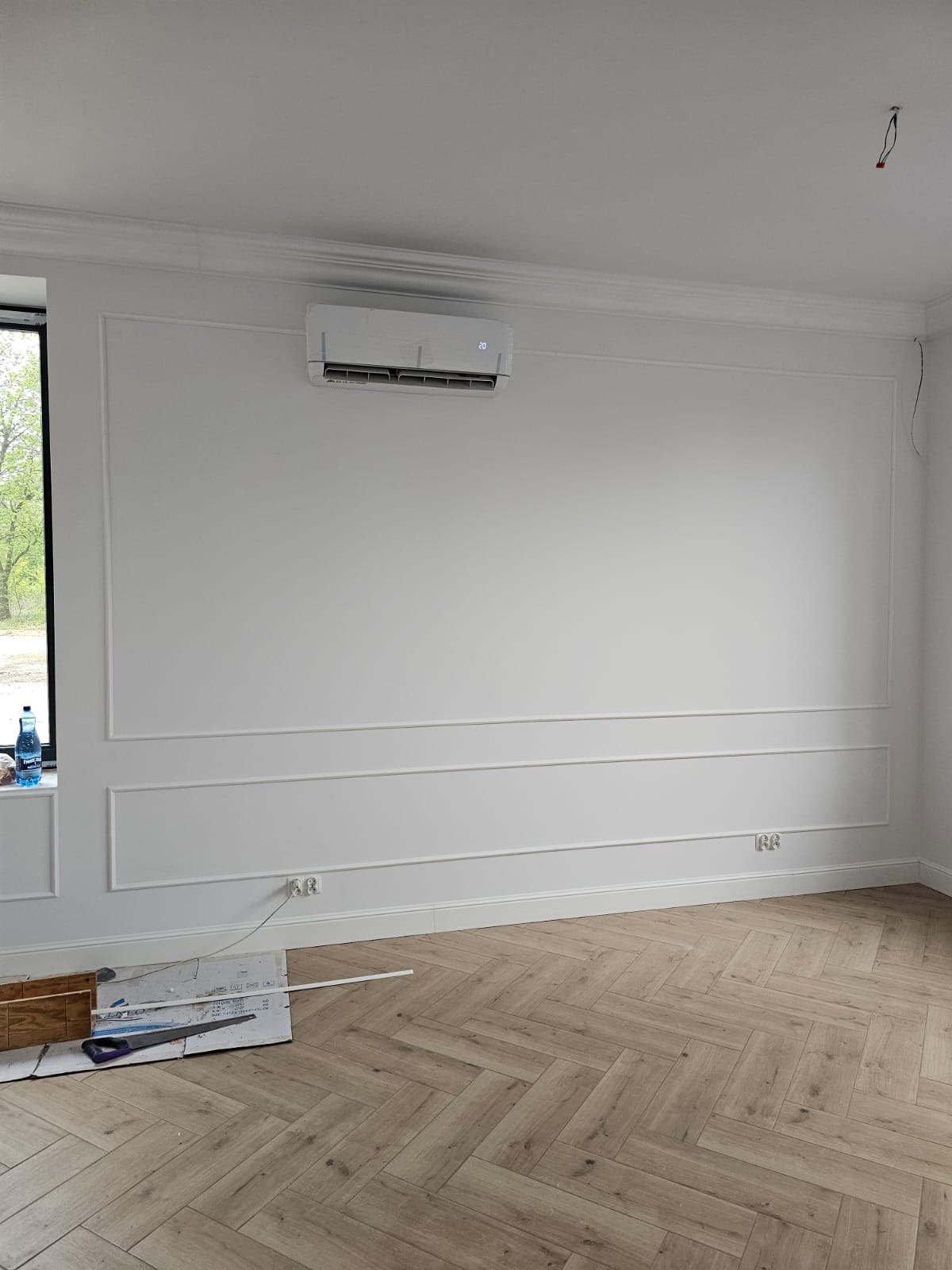 Pokój po remoncie z ułożonymi panelami podłogowymi w jodełkę, białe ściany z dekoracyjnymi listwami, klimatyzator na ścianie, widoczne narzędzia i materiały budowlane w rogu pokoju.