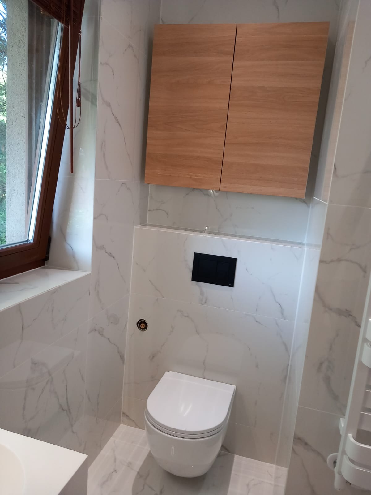 Nowoczesna łazienka z białą armaturą, marmurowymi płytkami i drewnianą szafką wiszącą nad toaletą.