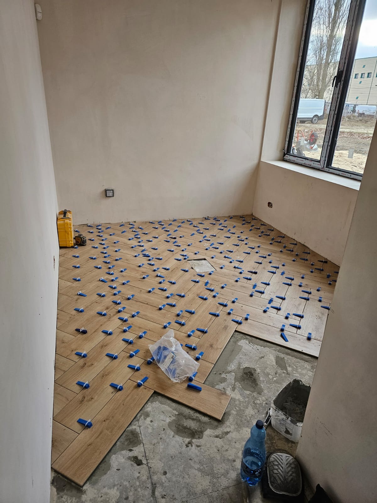 Układanie podłogi z płytek imitujących drewno w jodełkę, z użyciem niebieskich krzyżyków dystansowych; widoczne narzędzia i materiały budowlane, surowe ściany przed wykończeniem.