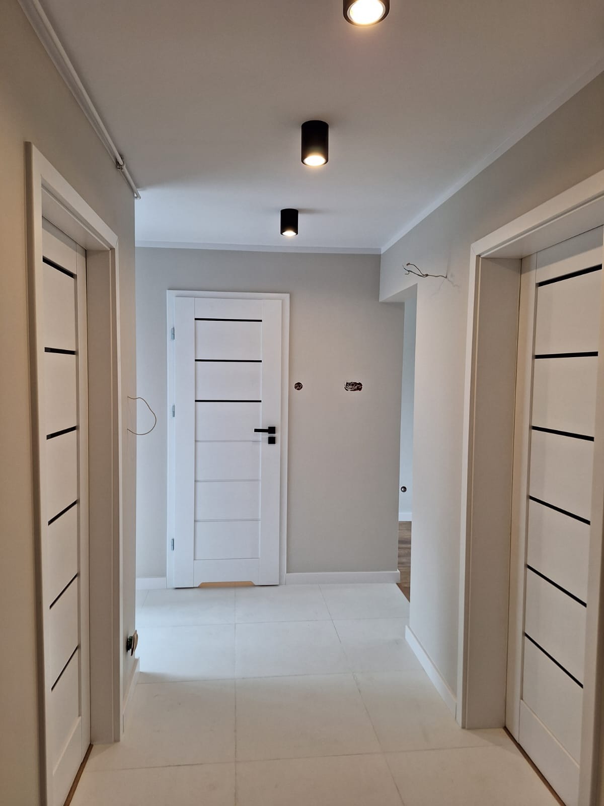 Korytarz w jasnych barwach z białymi drzwiami z czarnymi akcentami i kwadratowymi płytkami podłogowymi, oświetlony trzema czarnymi lampami sufitowymi.