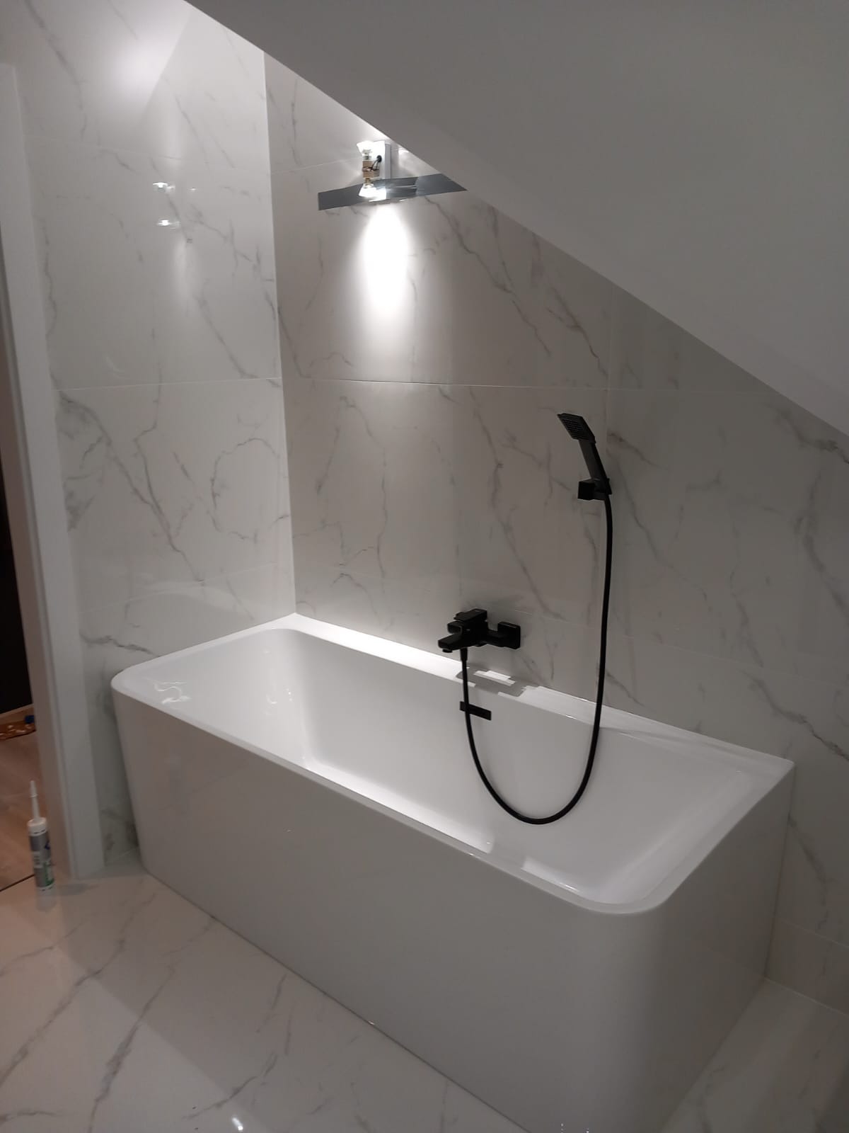 Biała, prostokątna wanna z czarną armaturą i słuchawką prysznicową na tle marmurowych płytek w łazience na poddaszu ze skosem.