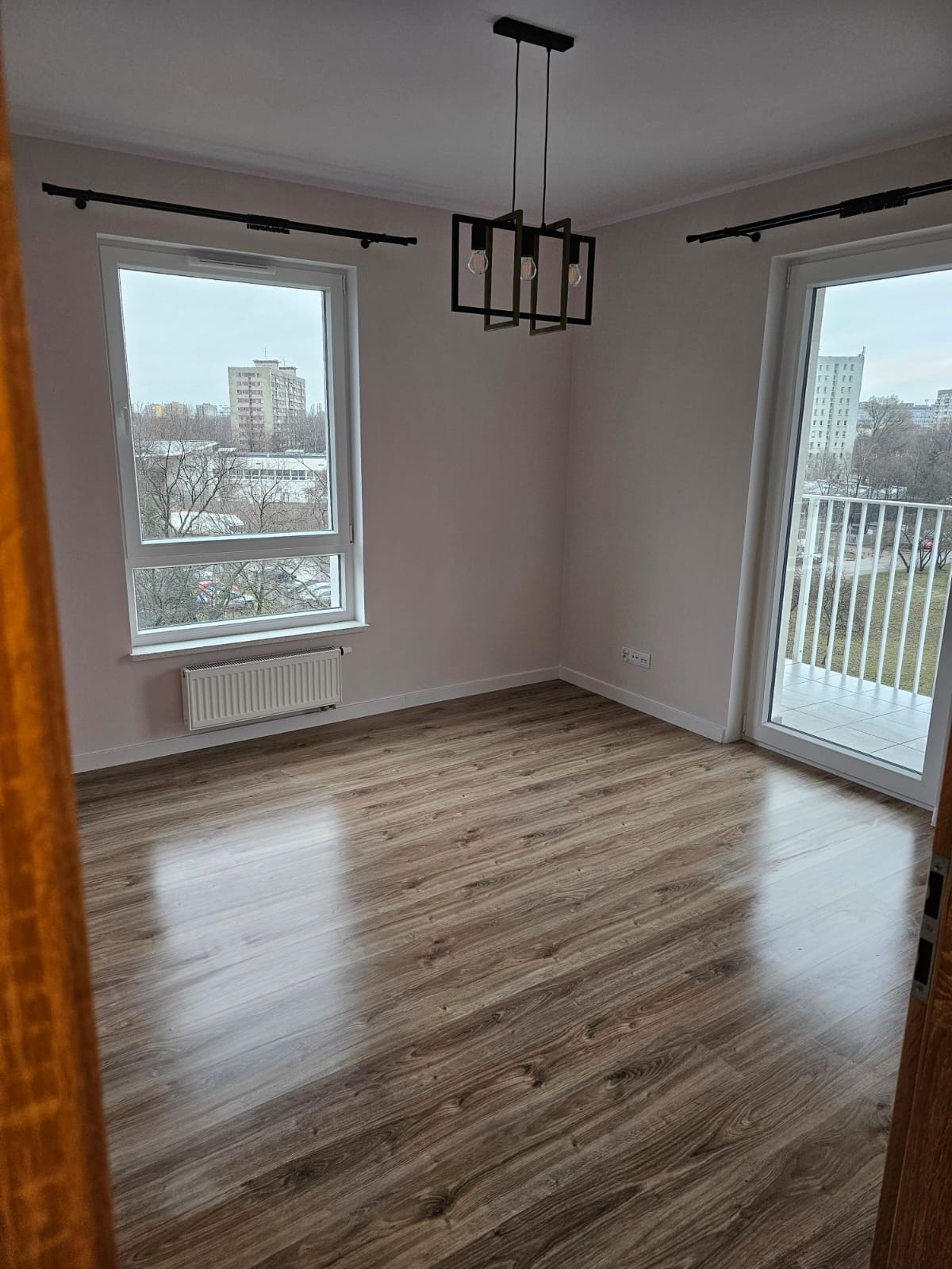 Pusty pokój po remoncie z panelami podłogowymi w odcieniach brązu, nowoczesną lampą wiszącą z trzema żarówkami w geometrycznej oprawie, oknem z widokiem na blok i balkonem z białą balustradą.