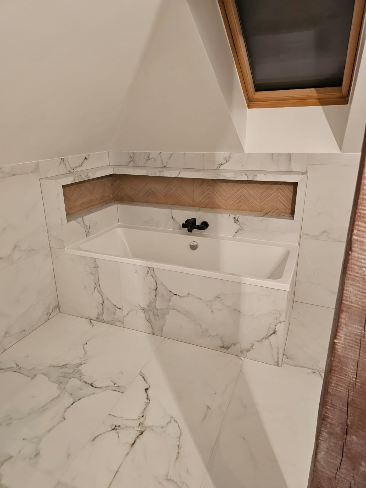 Nowoczesna łazienka na poddaszu z białą wanną obudowaną marmurowymi płytkami, czarna bateria, wnęka wykończona drewnem w jodełkę, okno dachowe