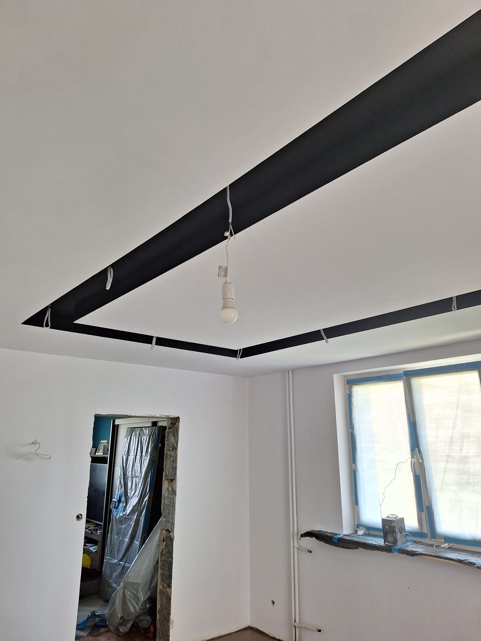 Wnętrze w trakcie remontu, widoczne czarne pasy na suficie tworzące ramę, ściany pomalowane na biało, okno zabezpieczone folią malarską, w tle widoczne drzwi z folią ochronną.