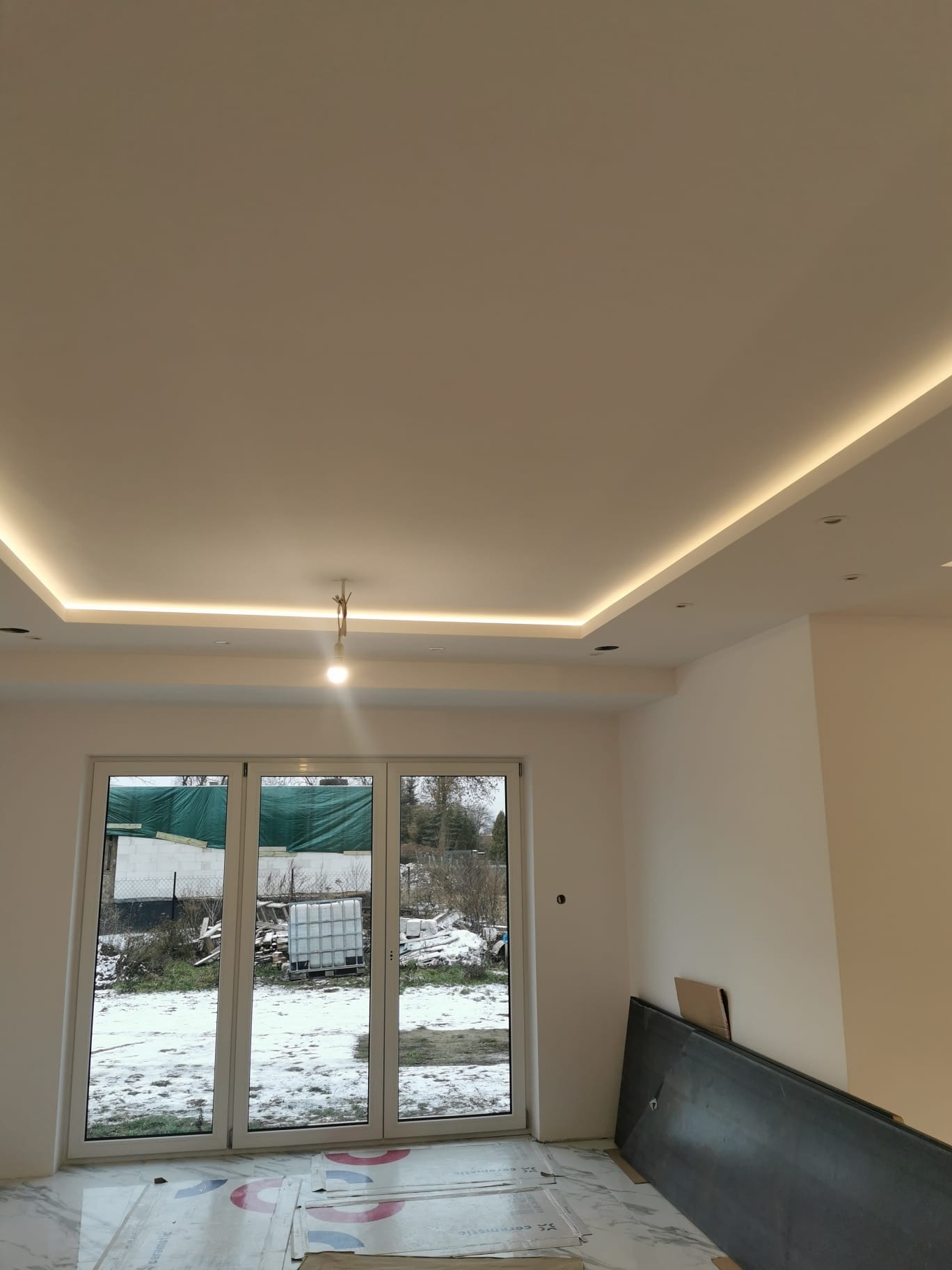 Nowoczesne oświetlenie sufitowe LED w świeżo wyremontowanym pokoju w Kobyłce, widok na okno z zimowym krajobrazem, panele podłogowe częściowo przykryte folią ochronną.