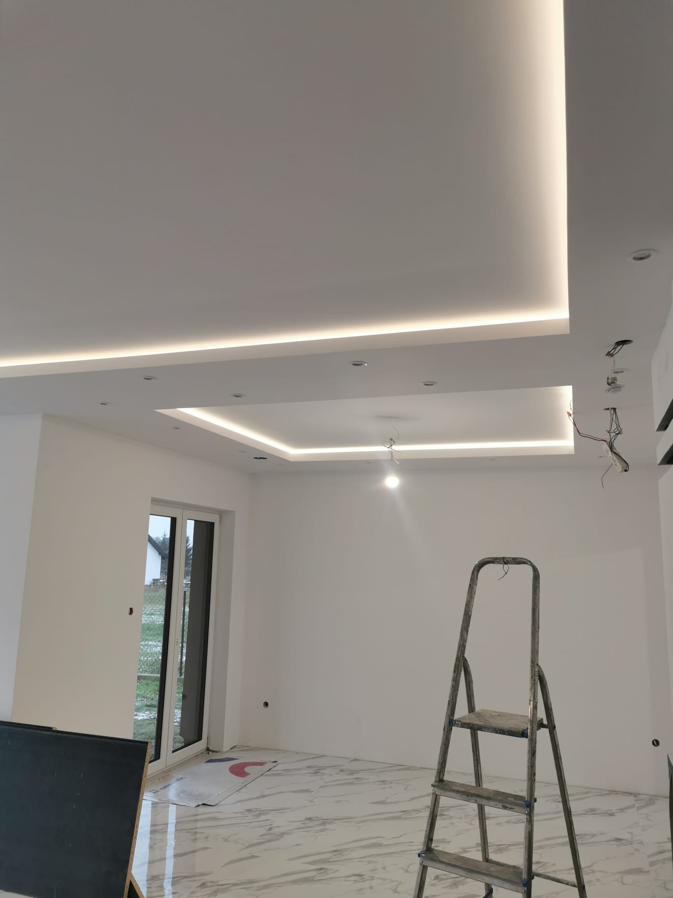 Wnętrze w trakcie wykańczania z podwieszonym sufitem i oświetleniem LED, marmurową podłogą, aluminiową drabiną i wiszącymi przewodami elektrycznymi.