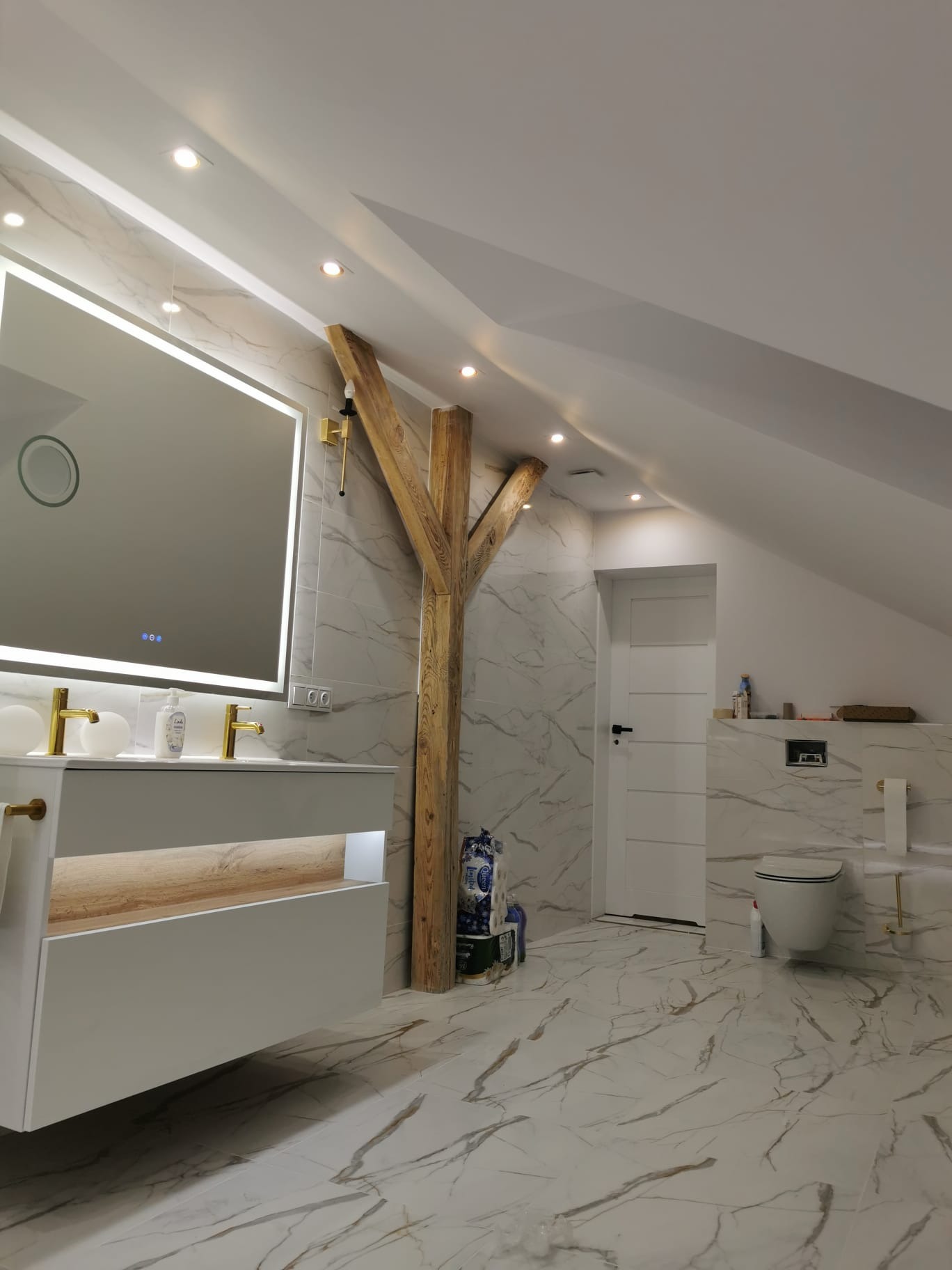 Jasna łazienka na poddaszu z marmurowymi płytkami, drewnianą belką konstrukcyjną, podświetlanym lustrem i złotą armaturą, widok na toaletę podwieszaną.