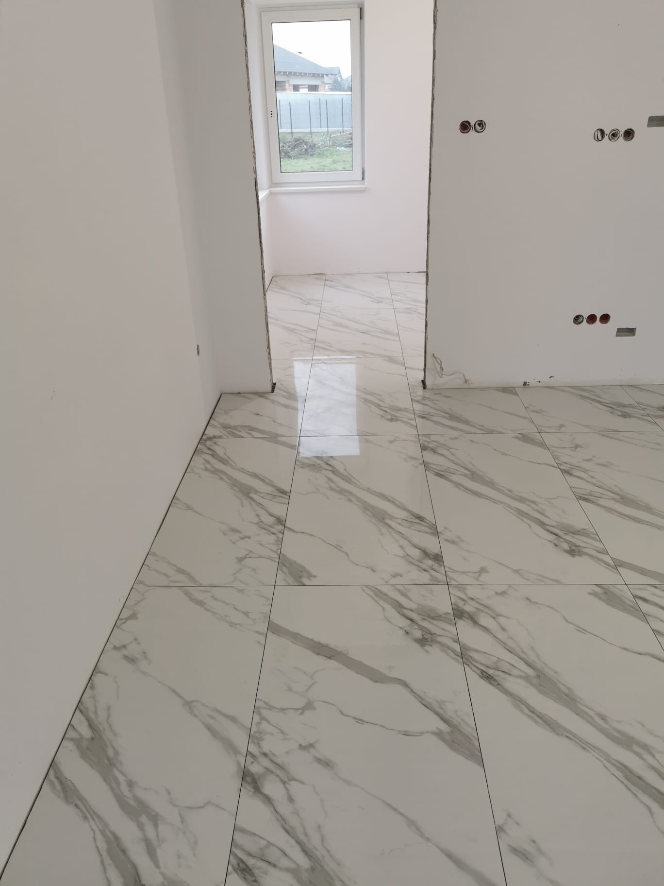 Białe płytki podłogowe z marmurkowym wzorem, świeżo położone w pustym pokoju z oknem, widoczne puszki elektryczne w ścianie.