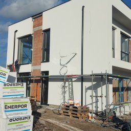 KARSBUD Karolina Zięcik - Dwukondygnacyjny dom z białą elewacją w trakcie prac wykończeniowych, widoczne rusztowania, styropian ENERPOR i pracownik przy oknie. Częściowo otynkowany budynek.
