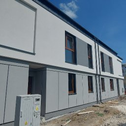 KARSBUD Karolina Zięcik - Nowoczesna elewacja szeregowca w odcieniach szarości i bieli. Widoczny fragment ulicy w budowie i zbiornik na zaprawę. Ujęcie z dołu, podkreślające geometrię budynku.