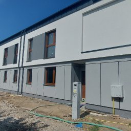 KARSBUD Karolina Zięcik - Nowoczesny dom w zabudowie szeregowej z szarą elewacją i drewnianymi oknami. Widoczne elementy instalacji zewnętrznej. W tle inne budynki i błękitne niebo.