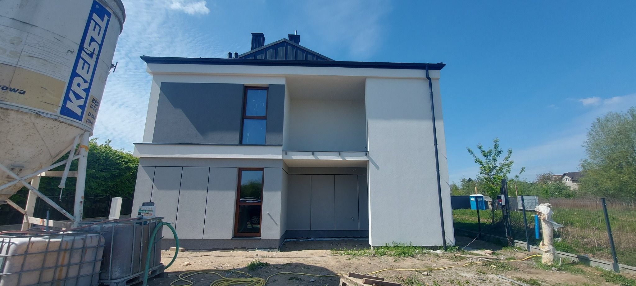 Nowoczesny dom z szarą elewacją i dużymi oknami, widziany z boku. Na pierwszym planie zbiornik z materiałami budowlanymi, sugerujący trwające prace wykończeniowe.