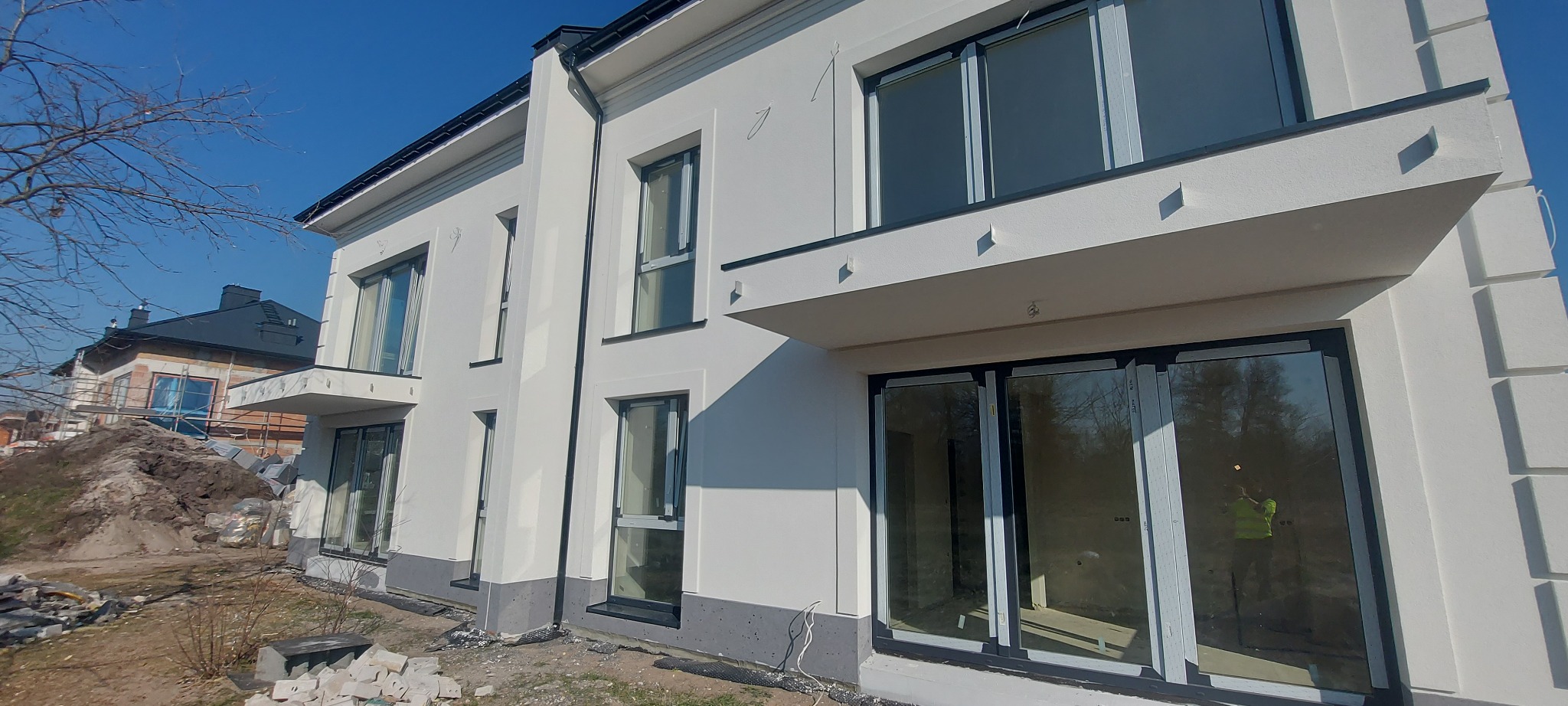Biała elewacja dwukondygnacyjnego budynku z czarnymi ramami okiennymi. W tle budowa innego domu i błękitne niebo. Widok z dołu.