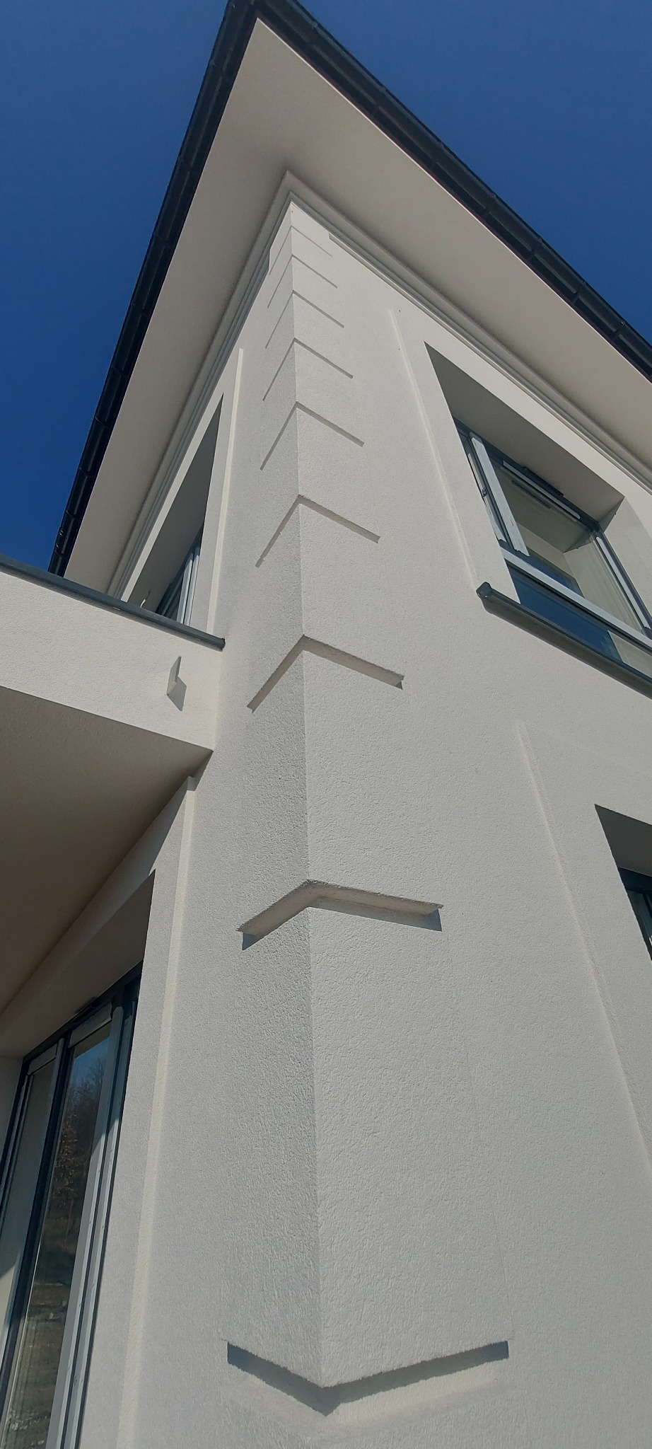 Biała elewacja budynku z dekoracyjnymi elementami w kształcie litery V, widok z dołu w słoneczny dzień. Tekstura tynku barankowego. Okna z szarymi ramami.