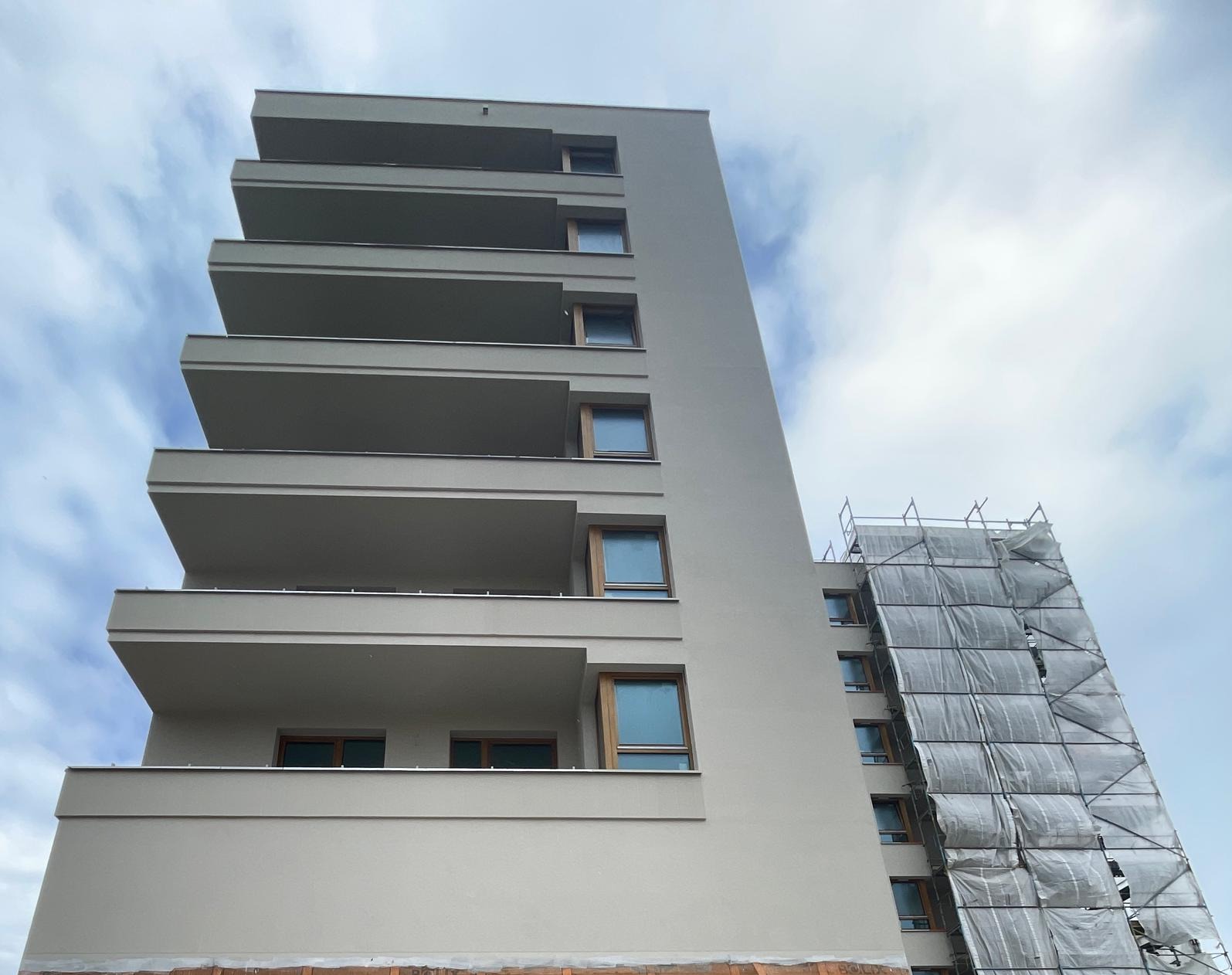 Nowoczesny, wysoki budynek z balkonami, częściowo zakryty rusztowaniem. Jasna elewacja, drewniane ramy okienne. Ujęcie z dołu, widoczne błękitne niebo.