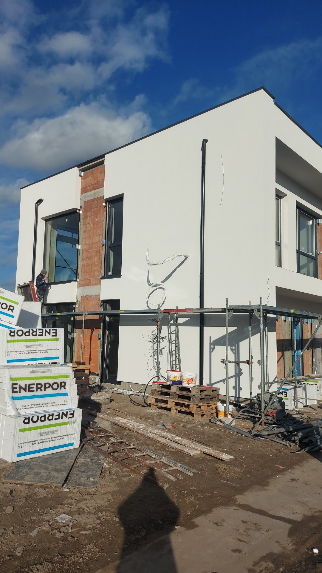 Dwukondygnacyjny dom z białą elewacją w trakcie prac wykończeniowych, widoczne rusztowania, styropian ENERPOR i pracownik przy oknie. Częściowo otynkowany budynek.