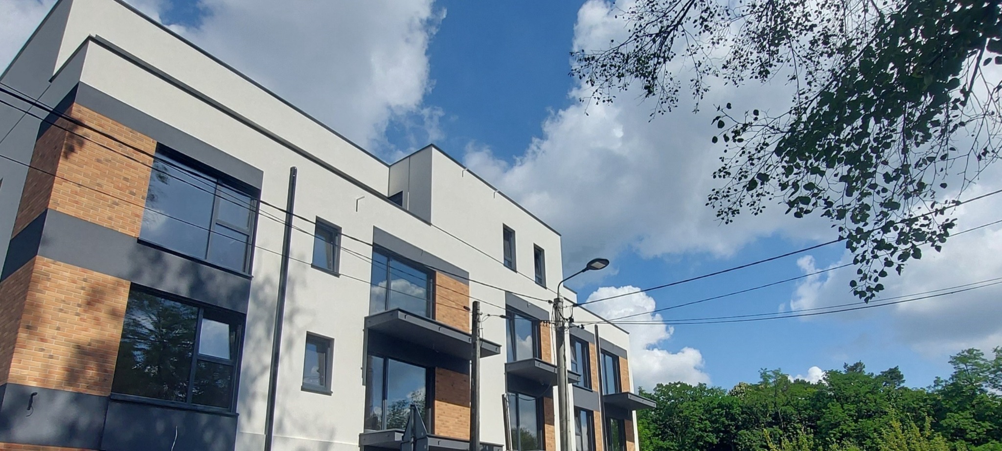 Nowoczesny budynek z elewacją w odcieniach bieli, szarości i cegły, z balkonami i dużymi oknami, na tle błękitnego nieba z chmurami.
