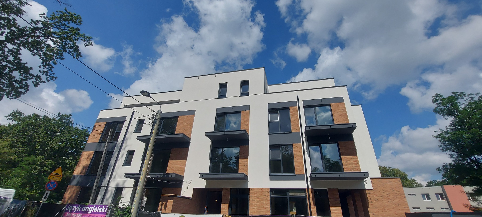 Nowoczesny budynek wielorodzinny z elementami cegły klinkierowej i białym tynkiem, balkony z szarymi balustradami, na tle błękitnego nieba z chmurami.