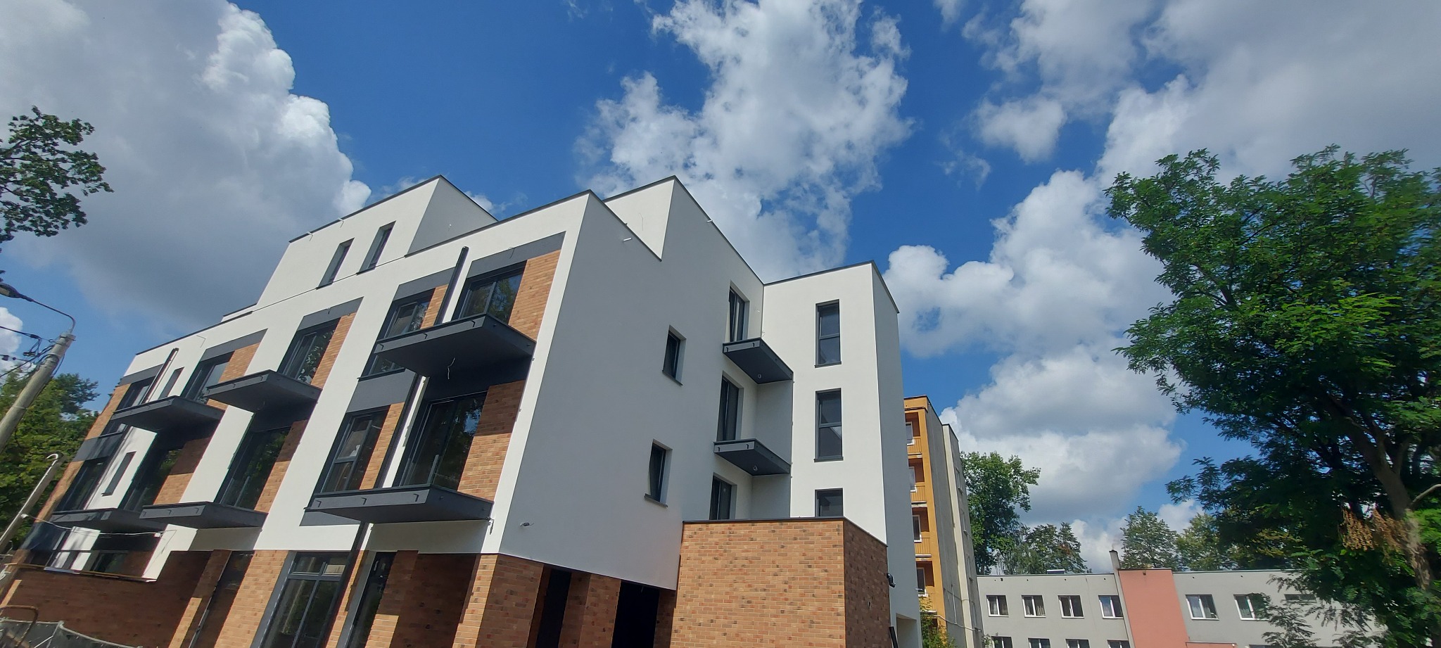 Nowoczesny budynek wielorodzinny z białymi ścianami i ceglanymi elementami, balkony z szarymi balustradami na tle błękitnego nieba z chmurami.