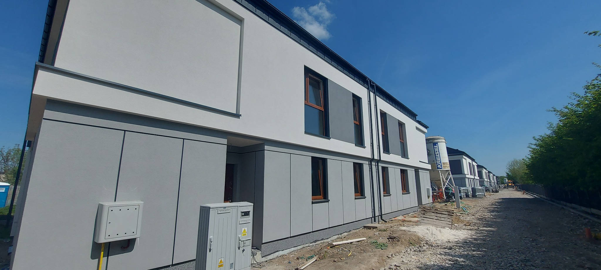 Nowoczesna elewacja szeregowca w odcieniach szarości i bieli. Widoczny fragment ulicy w budowie i zbiornik na zaprawę. Ujęcie z dołu, podkreślające geometrię budynku.