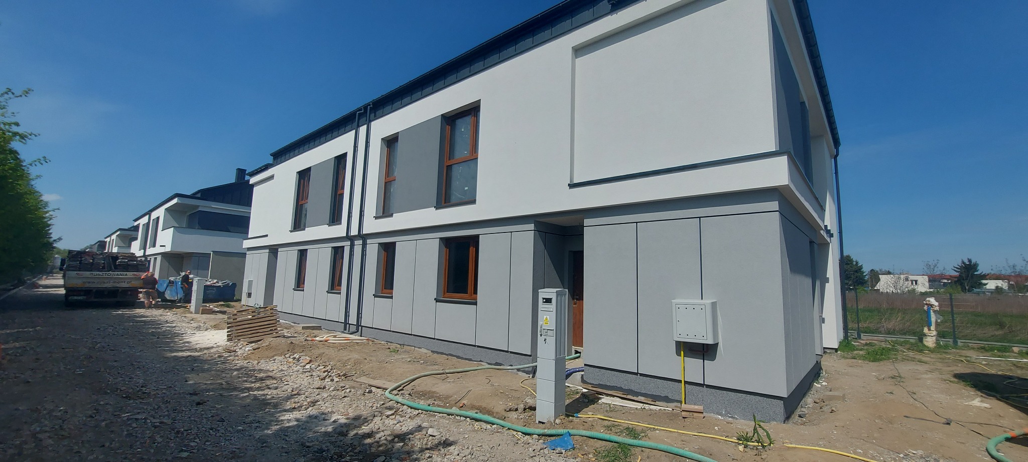 Nowoczesny dom w zabudowie szeregowej z szarą elewacją i drewnianymi oknami. Widoczne elementy instalacji zewnętrznej. W tle inne budynki i błękitne niebo.