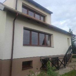 FIRMA HANDLOWO-USŁUGOWA DAMIAN GRODECKI - Elewacja domu po malowaniu: kremowa góra, brązowy dół. Widoczne okna z brązowymi ramami, rynna i schody z czarną barierką. Tło: zielona trawa i drzewa.