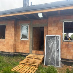 FIRMA HANDLOWO-USŁUGOWA DAMIAN GRODECKI - Budowa domu jednorodzinnego z czerwonej cegły. Widoczne okna wstawione w surowy mur, palety jako schody i pracownicy w oknach. Etap prac murarskich.