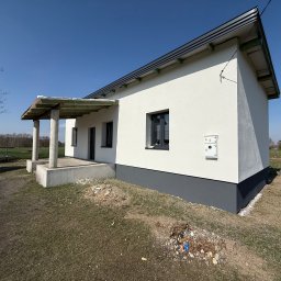 FIRMA HANDLOWO-USŁUGOWA DAMIAN GRODECKI - Biały dom z szarą podmurówką i zadaszonym wejściem, widok z boku na tle błękitnego nieba i zieleni. Widoczna skrzynka elektryczna na elewacji.