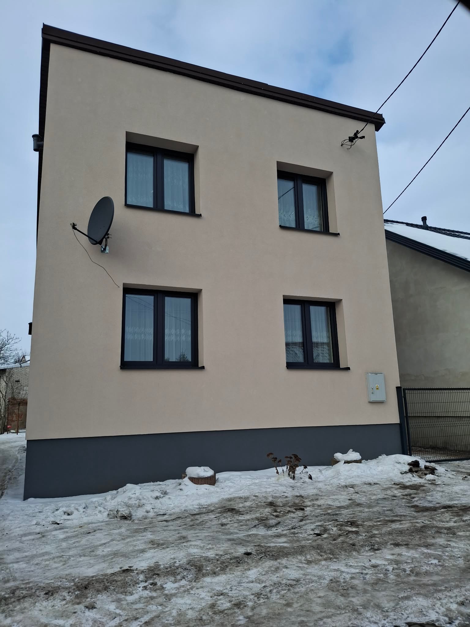Elewacja dwukondygnacyjnego domu po remoncie, z beżową fasadą, ciemnymi oknami i szarym cokołem. Widoczna antena satelitarna i skrzynka elektryczna. Zima, śnieg.