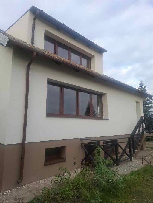 Elewacja domu po malowaniu: kremowa góra, brązowy dół. Widoczne okna z brązowymi ramami, rynna i schody z czarną barierką. Tło: zielona trawa i drzewa.