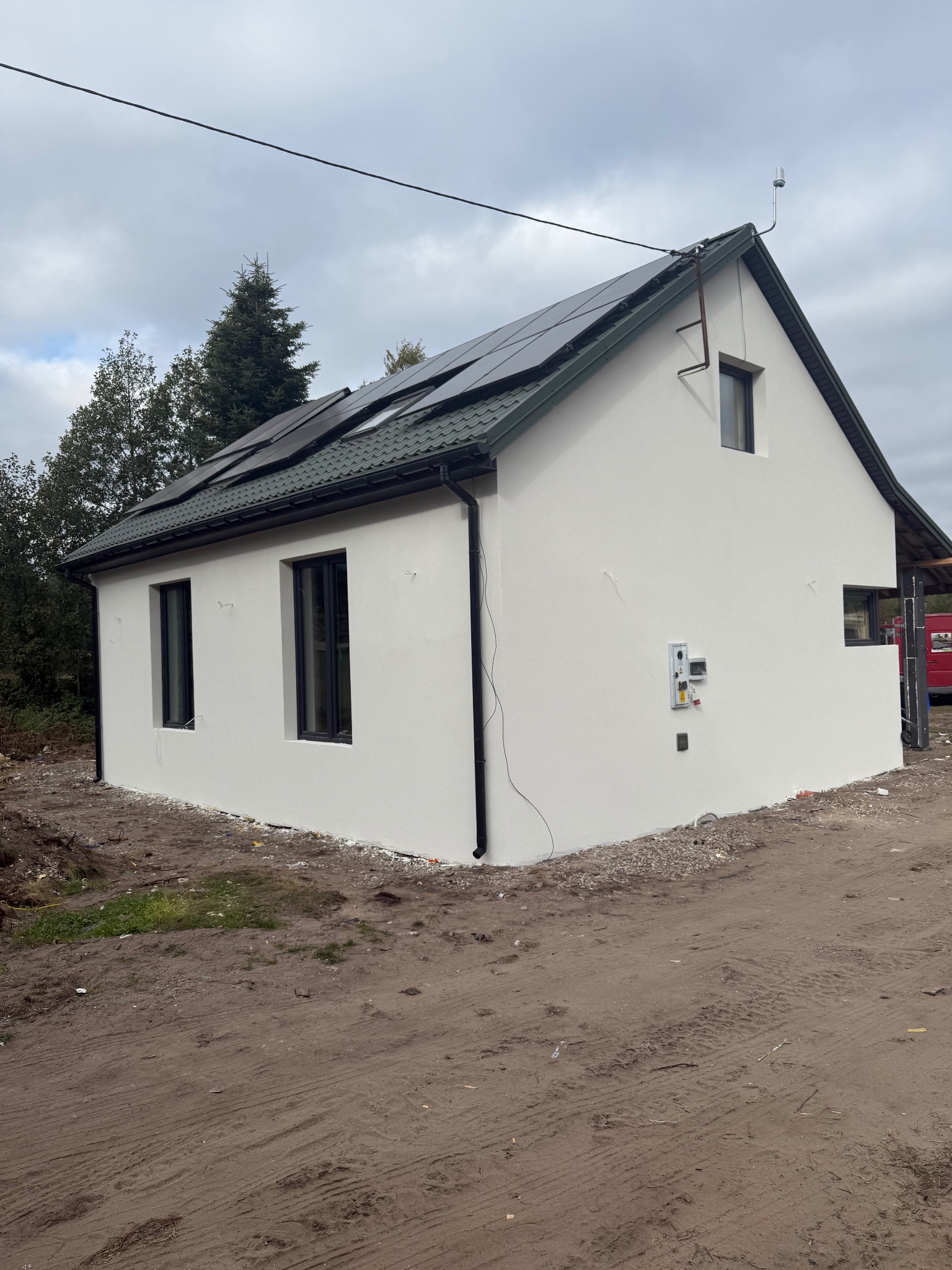 Otynkowany, biały budynek z panelami słonecznymi na dachu i czarnymi ramami okiennymi. Widoczne przyłącza elektryczne i teren wokół budynku.