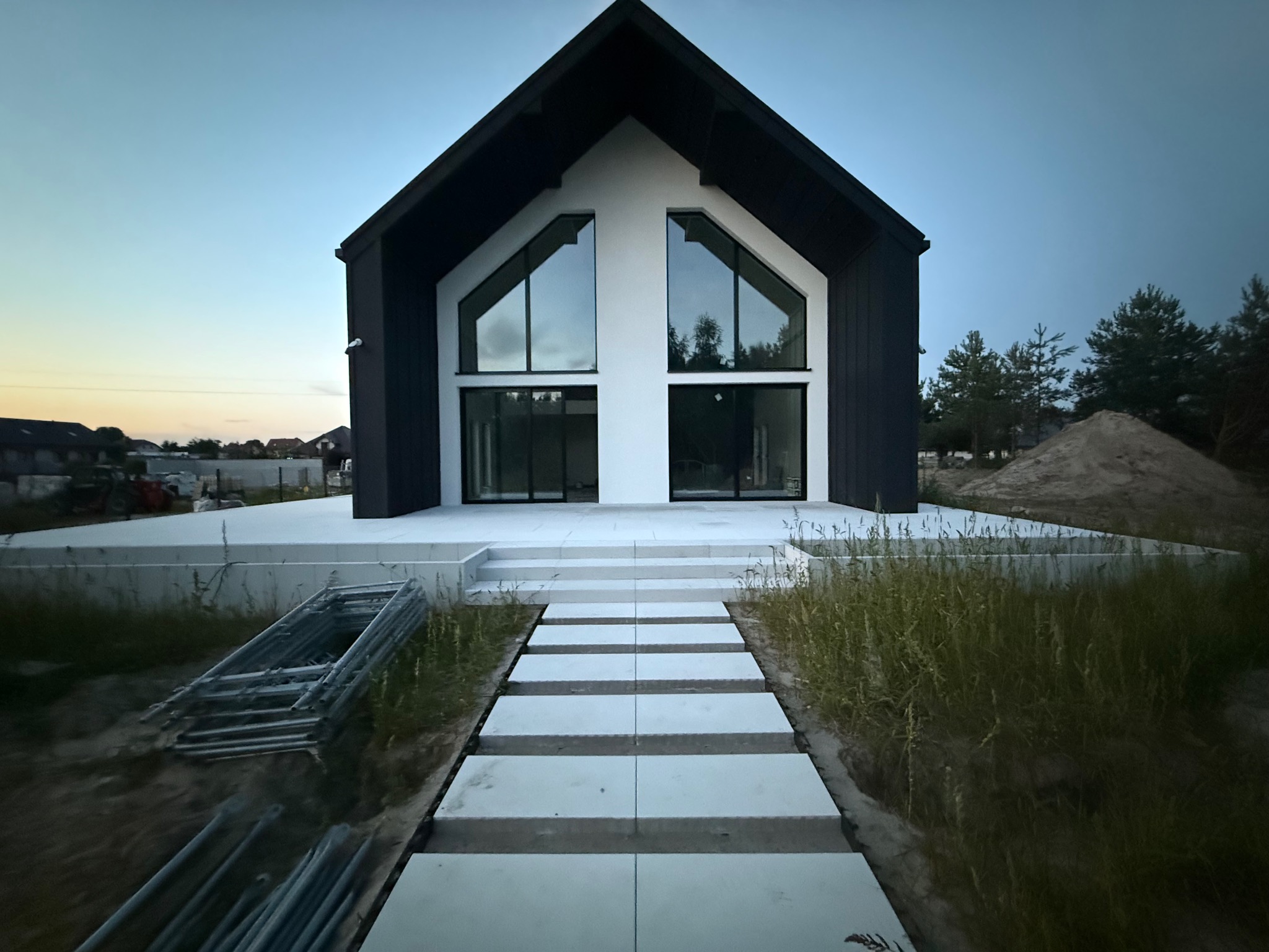 Nowoczesny dom z czarno-białą fasadą i dużymi oknami, widok z dołu na ścieżkę z betonowych płyt. W tle niebo o zmierzchu i elementy krajobrazu.
