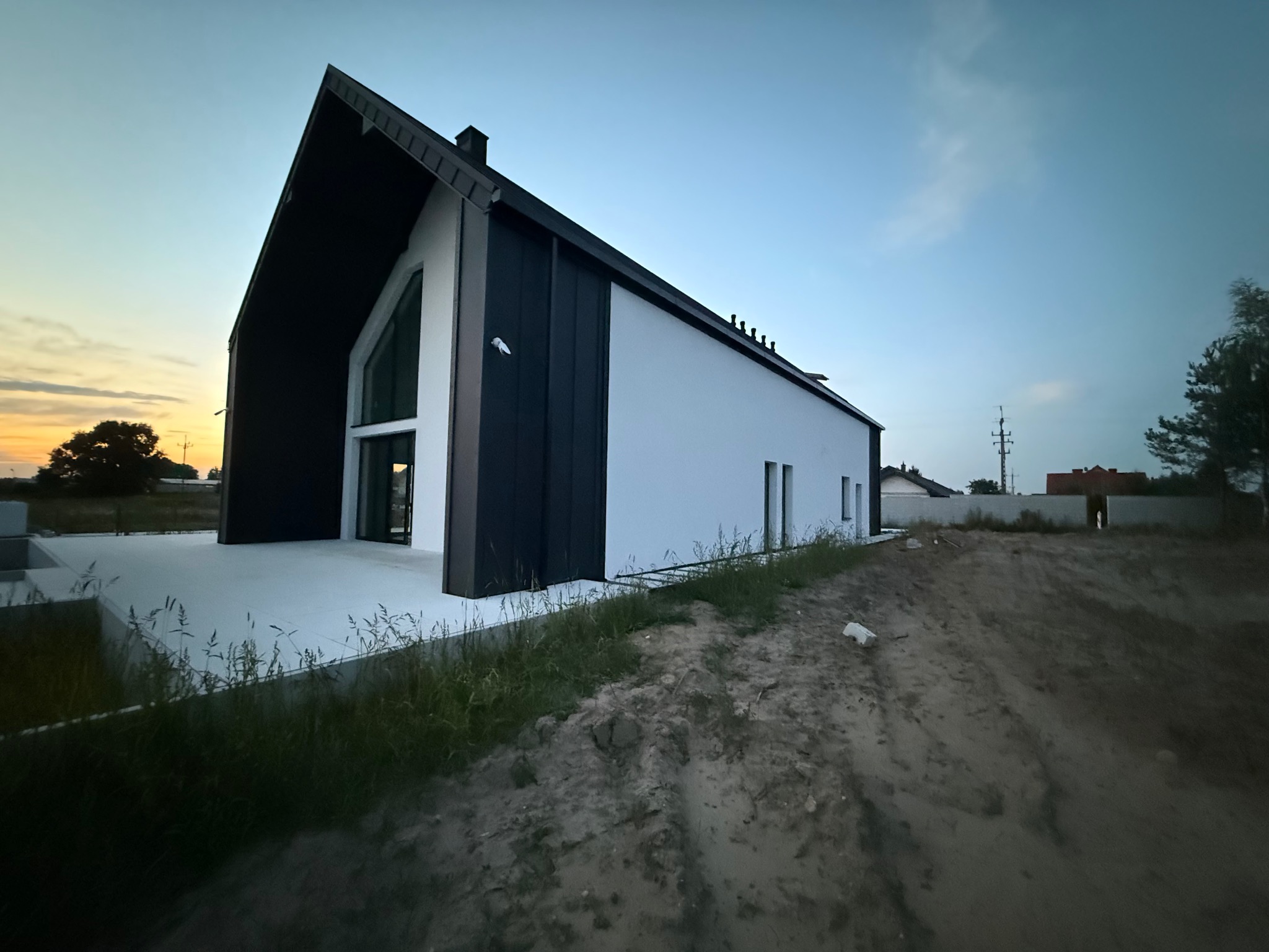 Nowoczesny dom jednorodzinny z białymi ścianami i czarnym dachem, widok z poziomu gruntu na tle błękitnego nieba o zmierzchu. Minimalistyczny design.