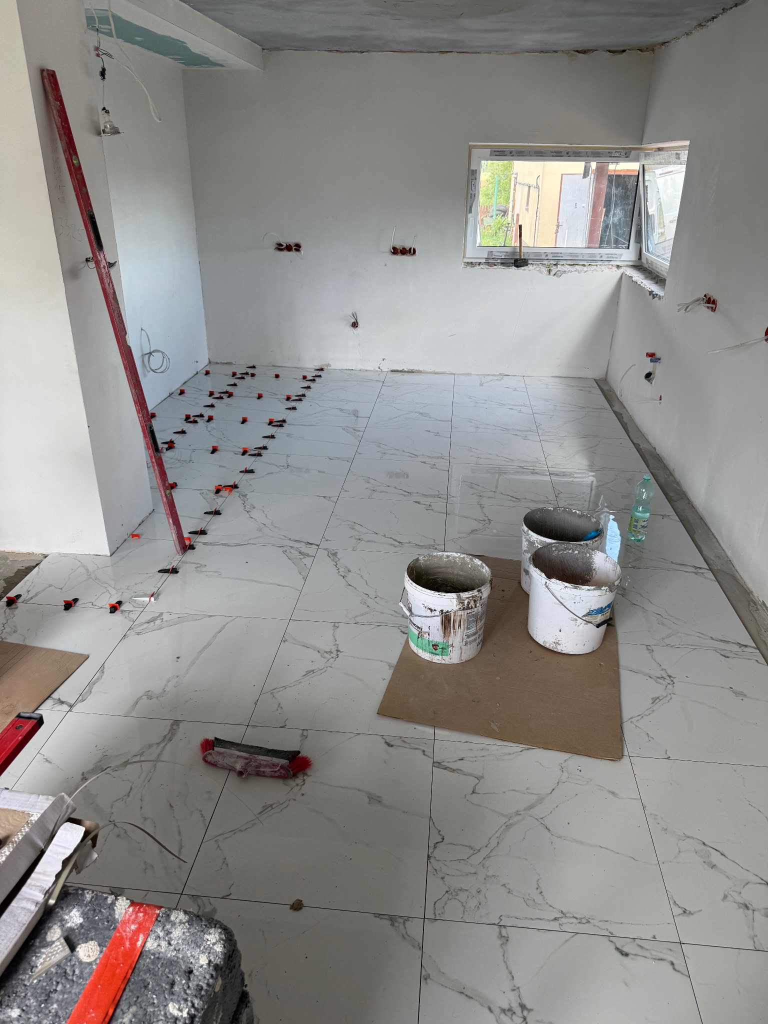 Układanie białych płytek imitujących marmur w pomieszczeniu z oknem. Widoczne poziomica, wiadra z zaprawą i krzyżyki dystansowe.