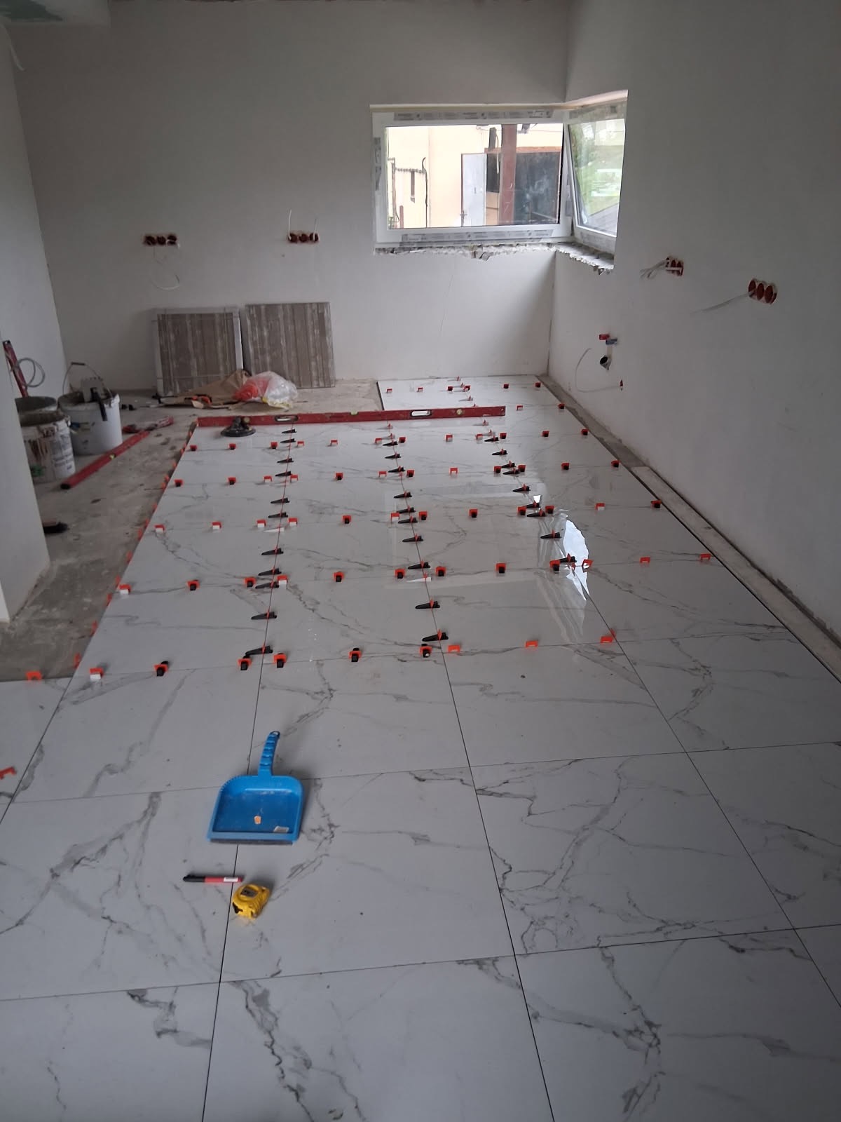 Układanie białych płytek podłogowych z marmurowym wzorem, z użyciem klipsów poziomujących. Widoczne narzędzia: miarka, poziomica, szufelka. Stan surowy, instalacja elektryczna.