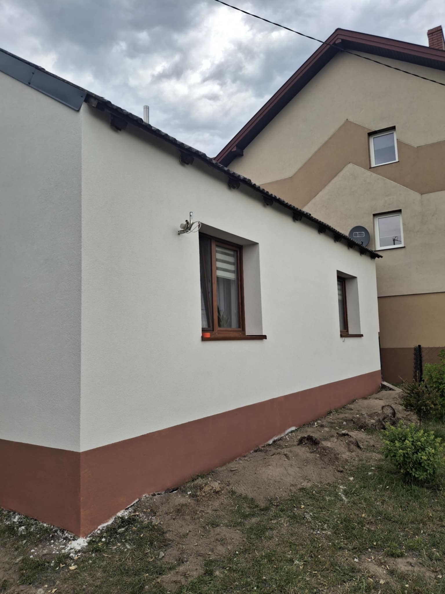 Odnowiona elewacja budynku z białym tynkiem i brązowym cokołem, widok z boku na tle sąsiedniego domu i pochmurnego nieba. Dwa okna z brązowymi ramami.