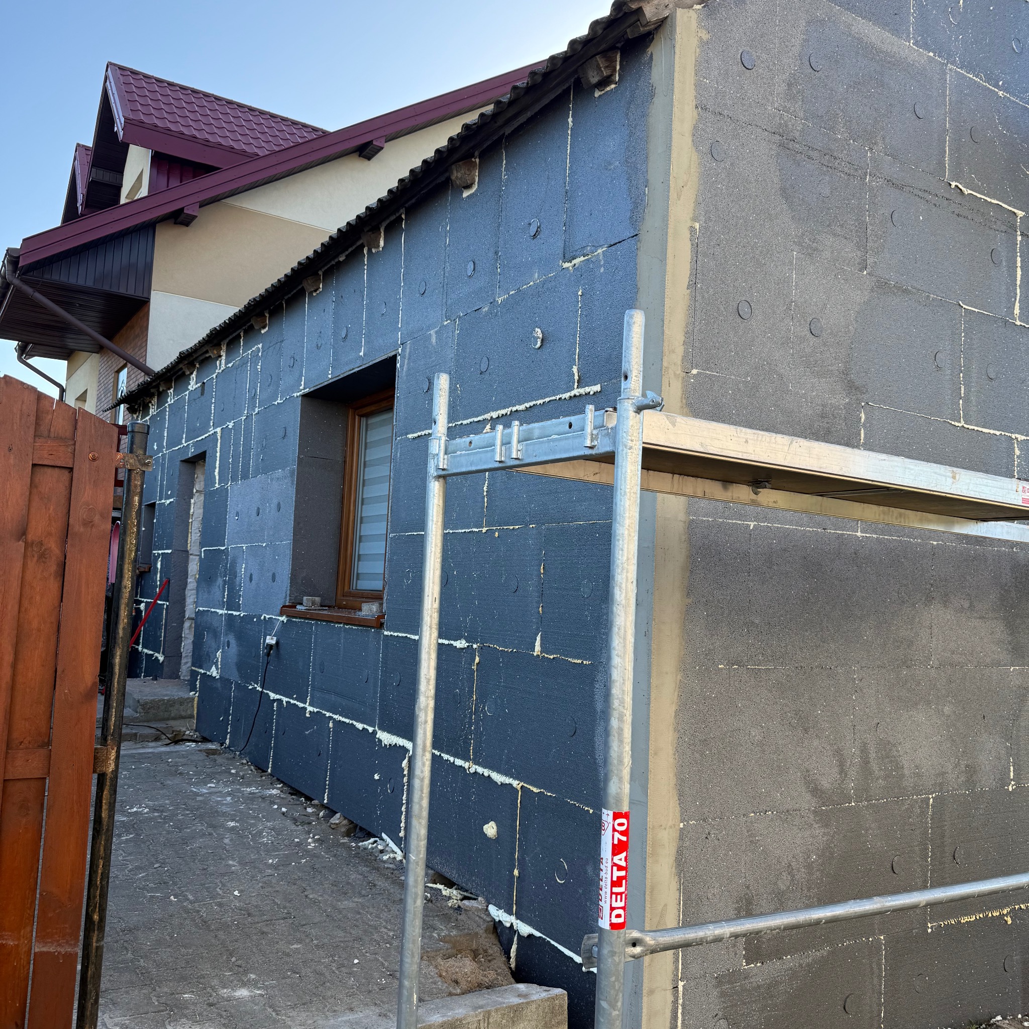 Ocieplanie ściany budynku styropianem grafitowym, widoczne rusztowanie i fragment domu z czerwoną dachówką. Etap prac przed nałożeniem tynku.